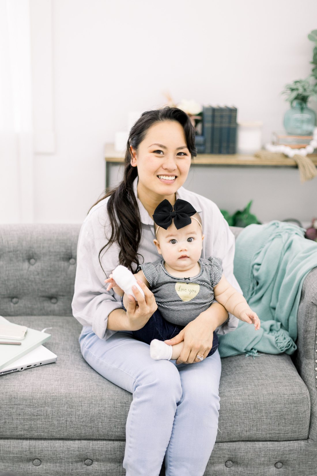 Serena Kretchmer smiling on a gray sofa, holding her baby, with a laptop and notebook beside her.